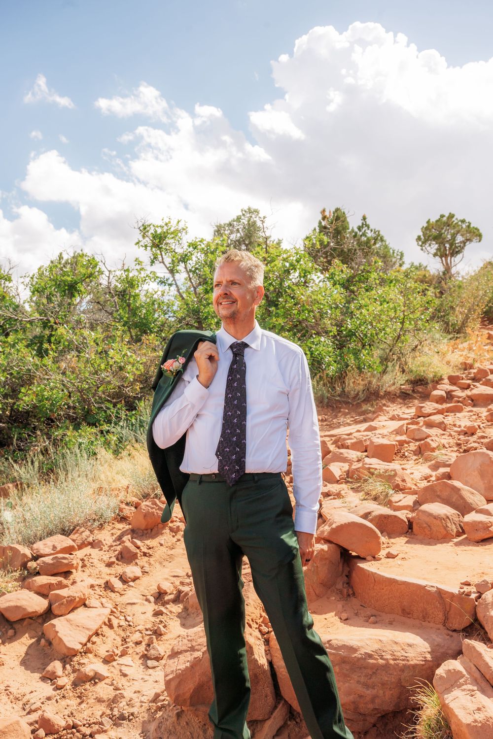 A businessman in a white shirt and tie carries his jacket over his shoulder while standing on a rocky red desert trail.