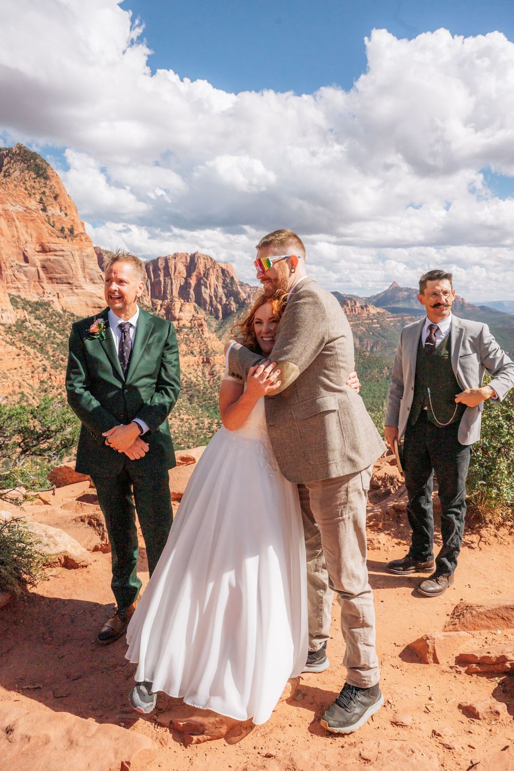 Couple embraces during outdoor wedding ceremony on red rock cliff in Zion National Park with guests and mountains behind.
