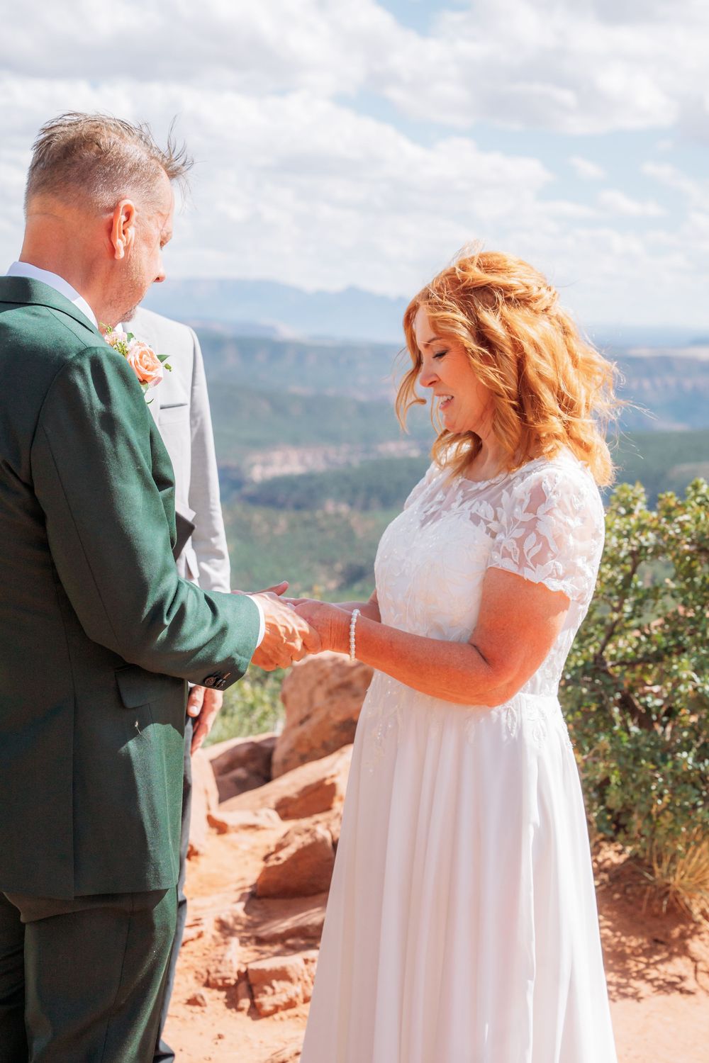 Couple exchanging rings during outdoor mountain wedding ceremony, bride in white lace dress, groom in green suit.