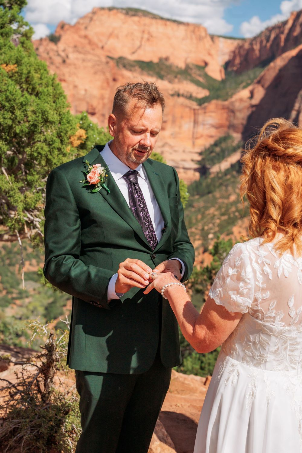 Groom in green suit places wedding ring on bride's finger with red rock canyon backdrop at Zion National Park.