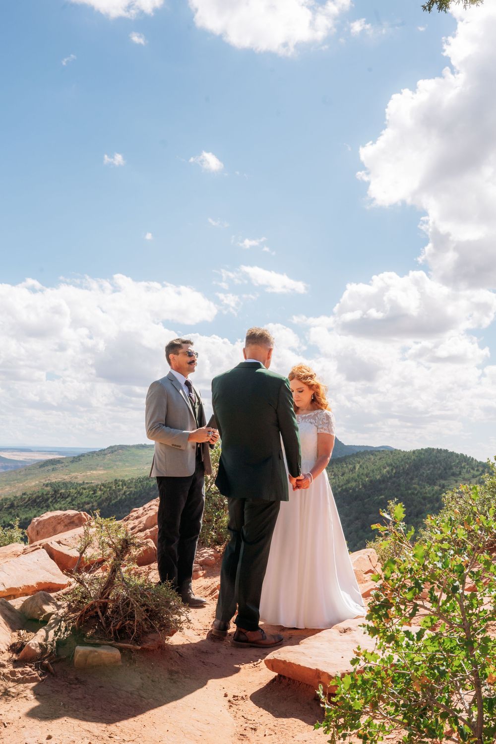 Couple exchanges vows with officiant on red rock summit overlooking lush green valley under cloudy sky.