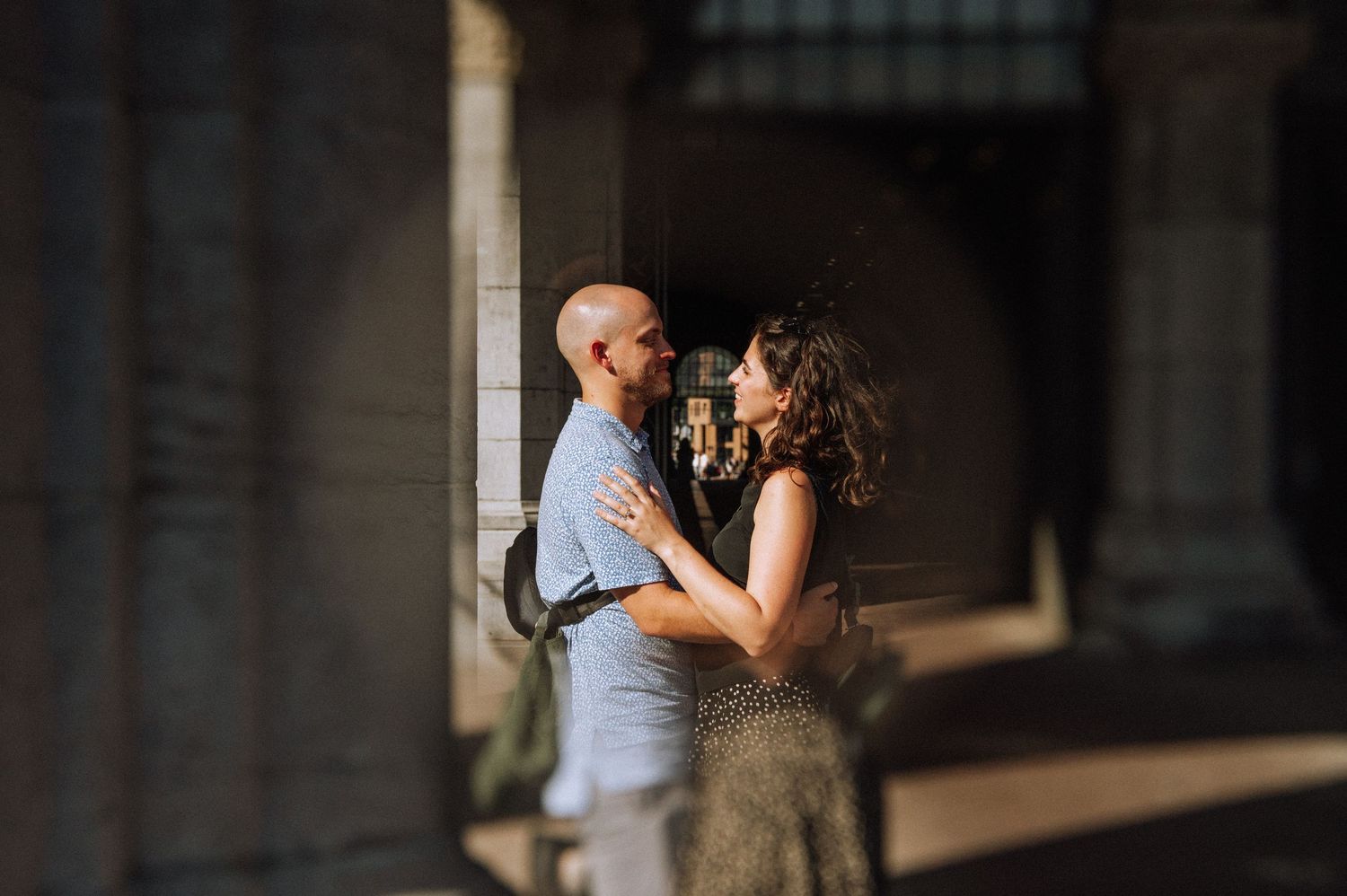 A couple embraces in a sunlit stone archway, the bald man in a blue shirt holding a curly-haired woman close in a warm, romantic moment.