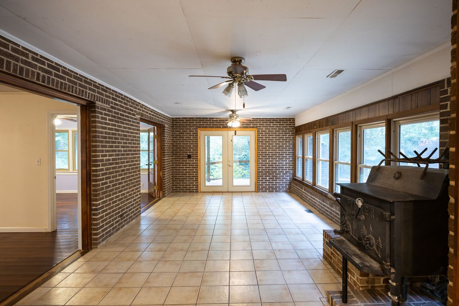 Spacious brick-walled living room with tile floors, ceiling fan, wood-burning stove, and French doors letting in natural light.