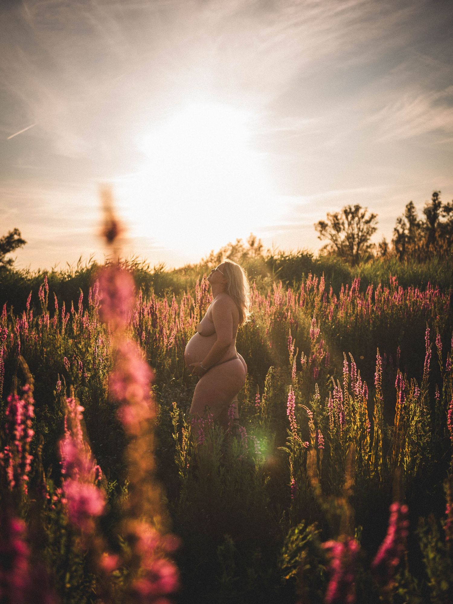 Pregnant woman standing in a field of pink wildflowers during golden sunset, glowing backlit maternity photography.