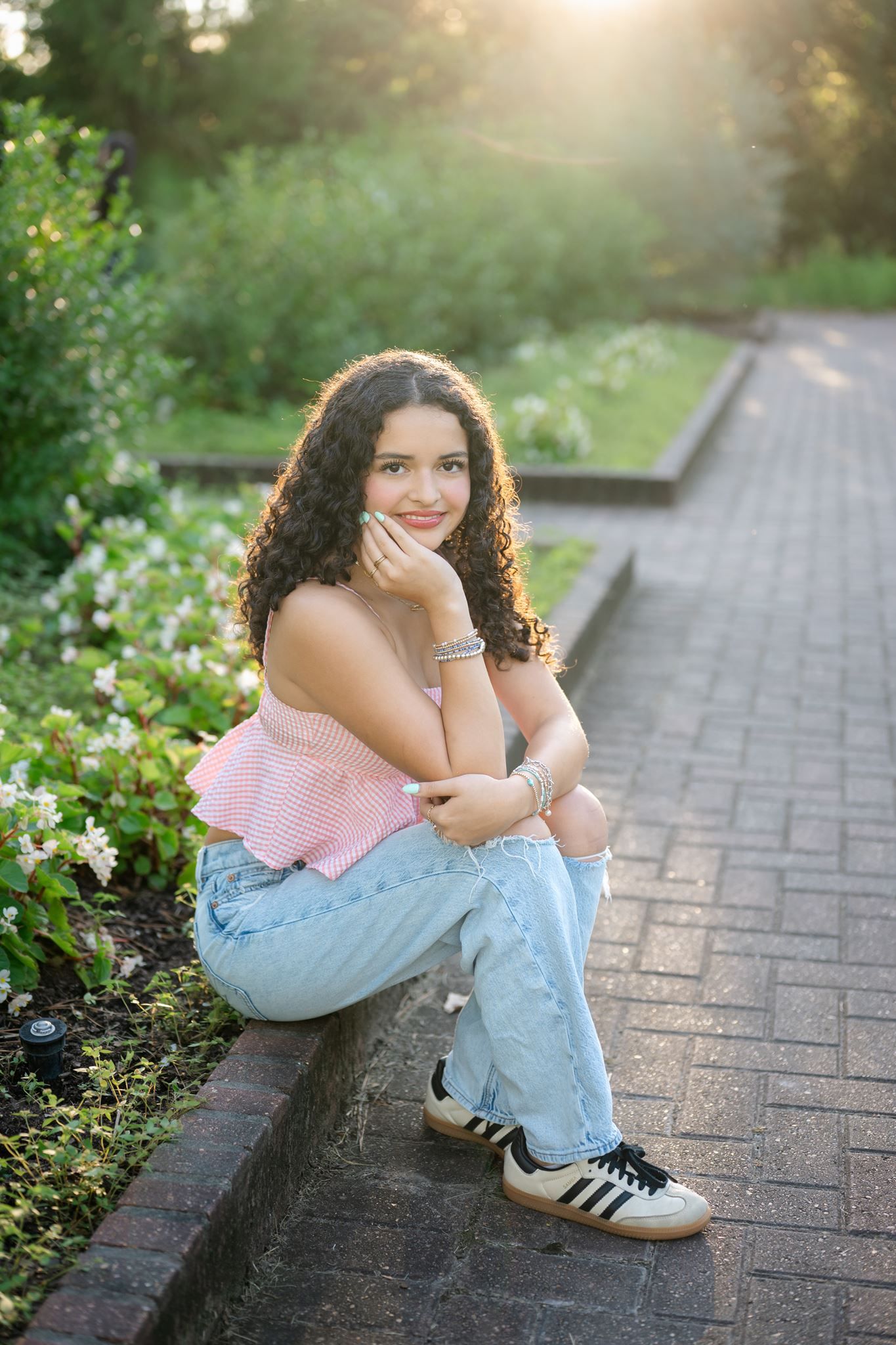 Senior girl with long curly hair in a pink top and light jeans sits on a brick garden ledge, smiling softly in warm golden sunlight.