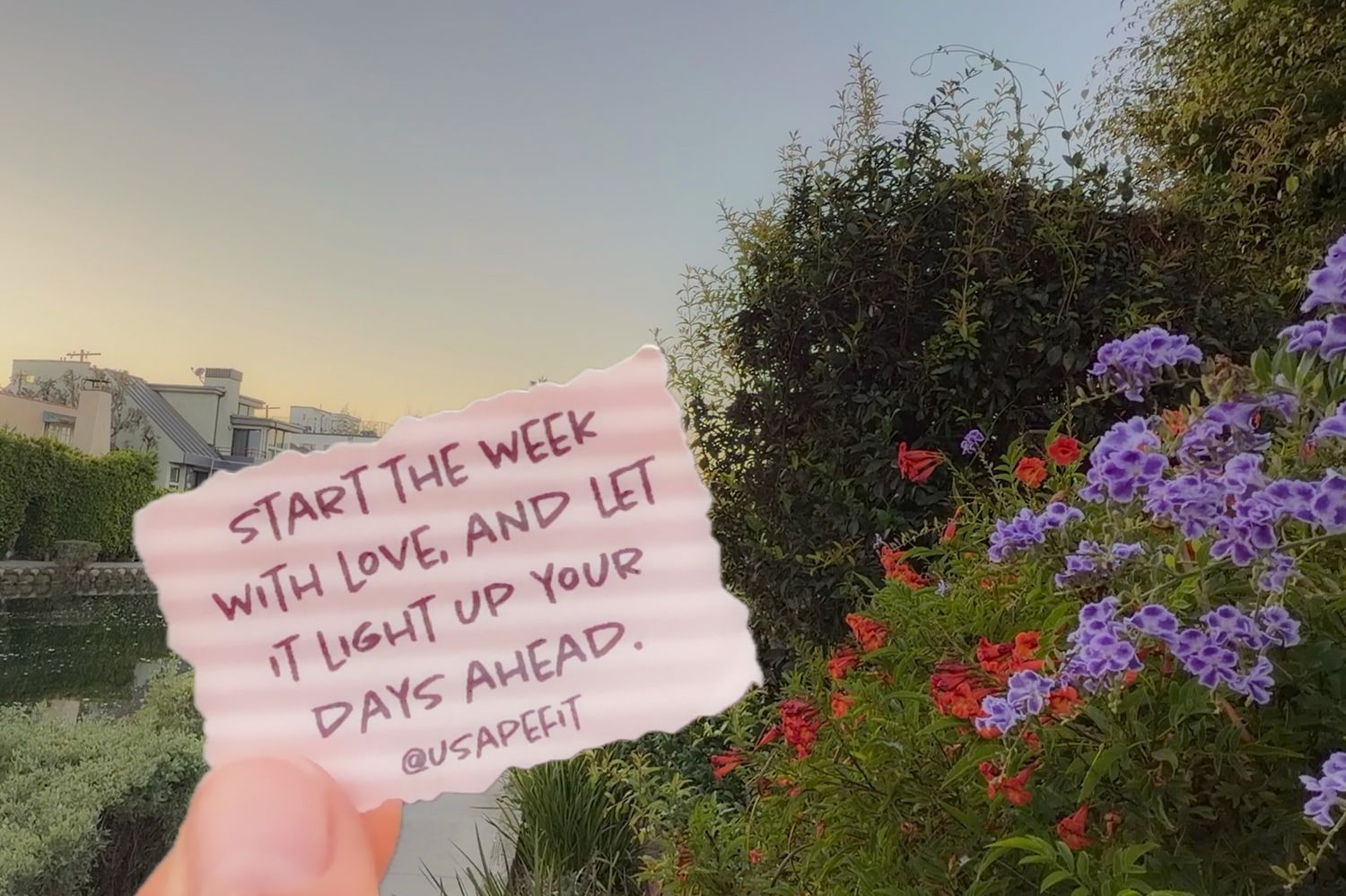 Hand holding a pink note reading "Start the week with love" before purple and red flowers, a canal, and rooftops at sunset.