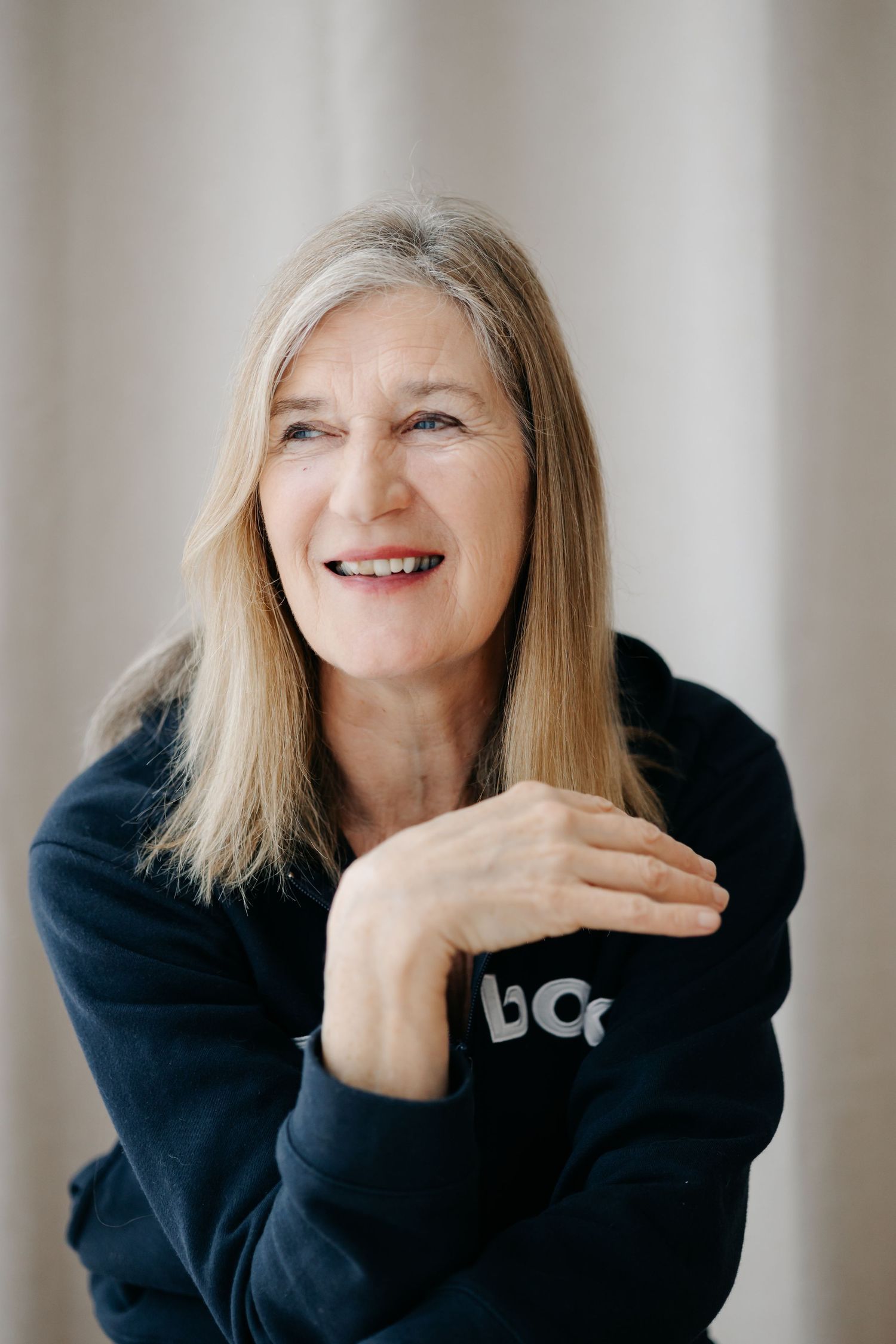 Smiling middle-aged woman with long blonde hair wearing a navy hoodie, posing with hands crossed in a bright room.