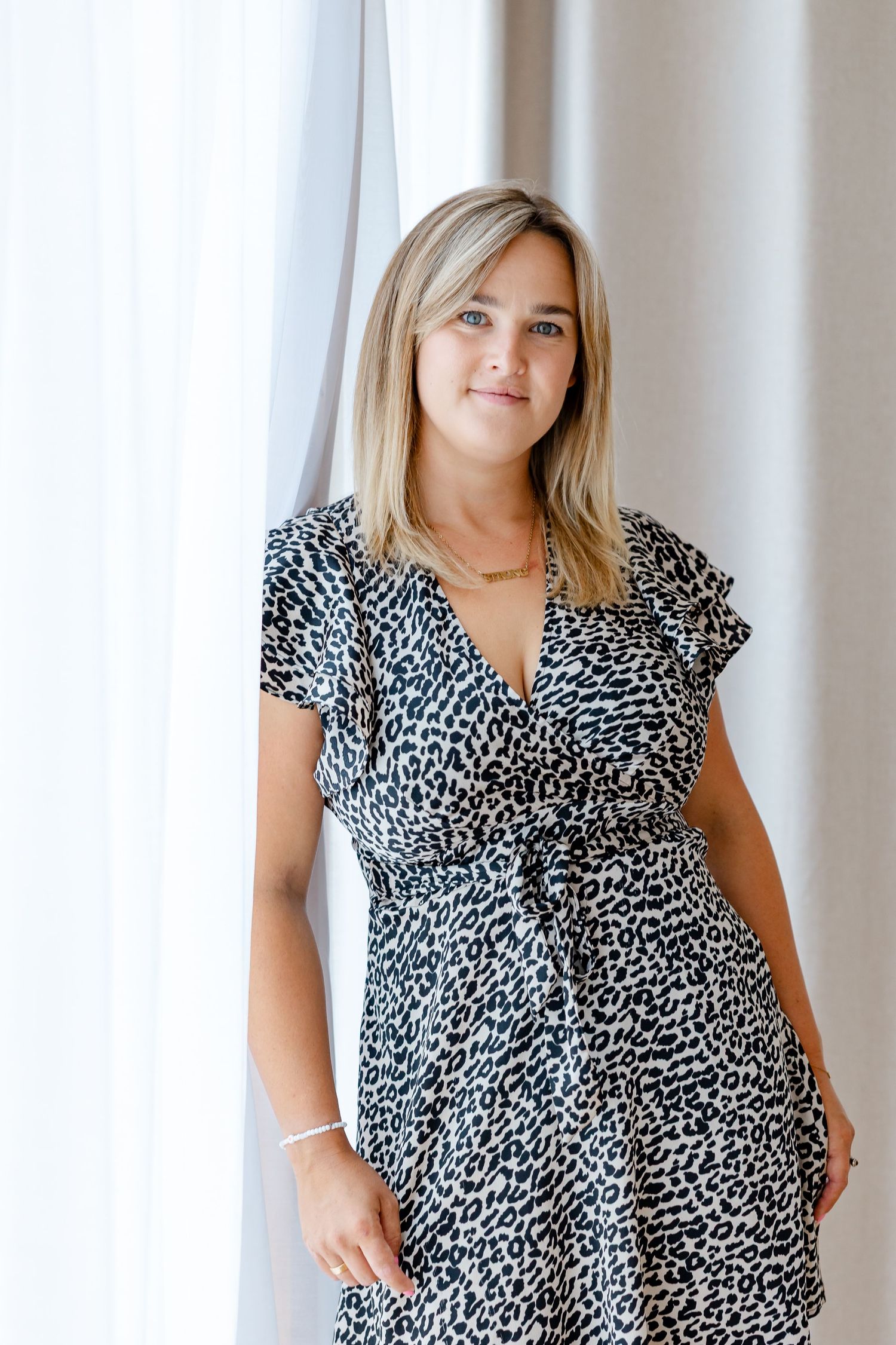 Blonde woman in leopard print wrap dress standing by sheer white curtains, smiling softly at camera.