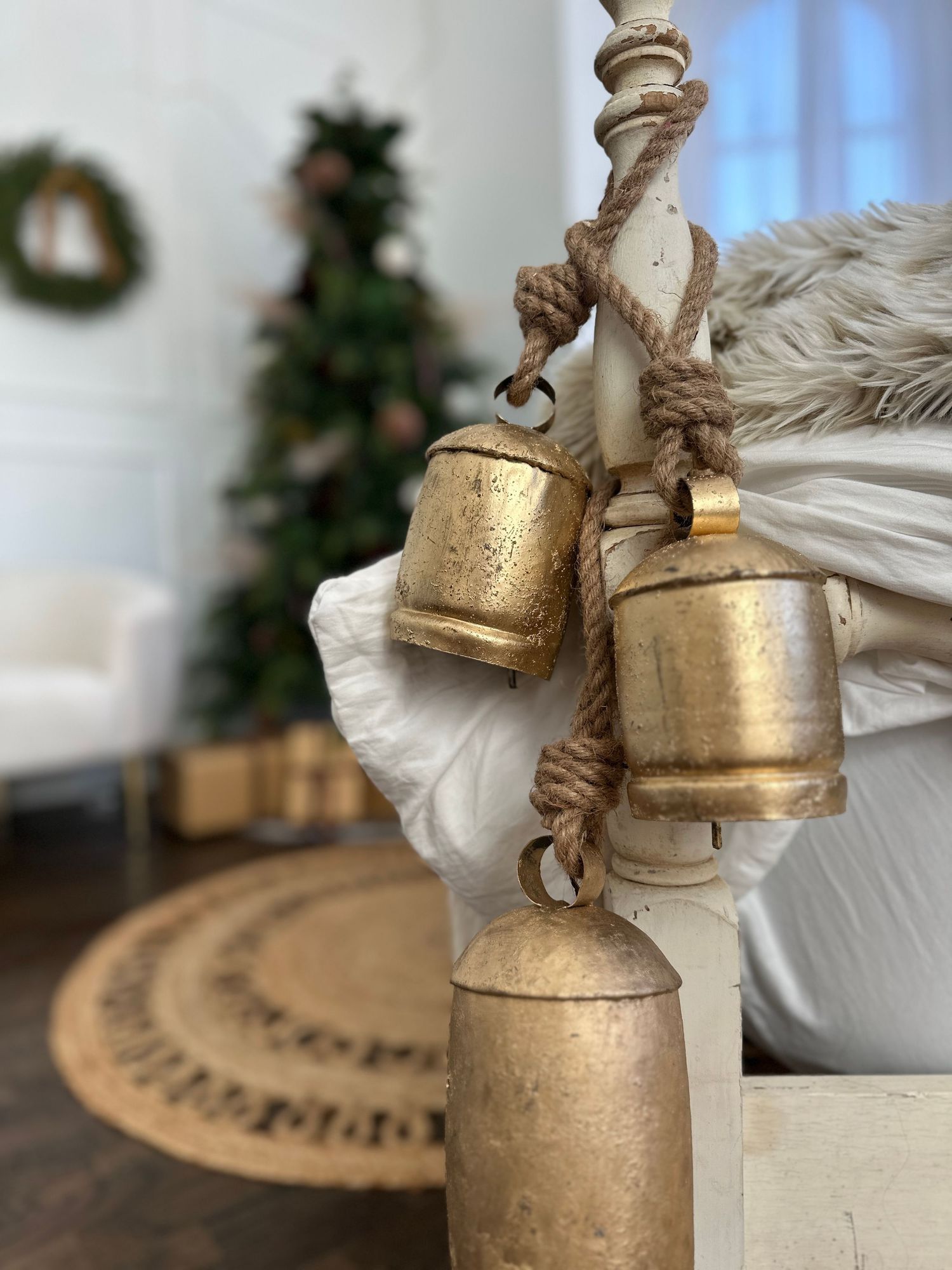 Three rustic gold bells hanging on a jute rope with a Christmas tree and wreath in the background.