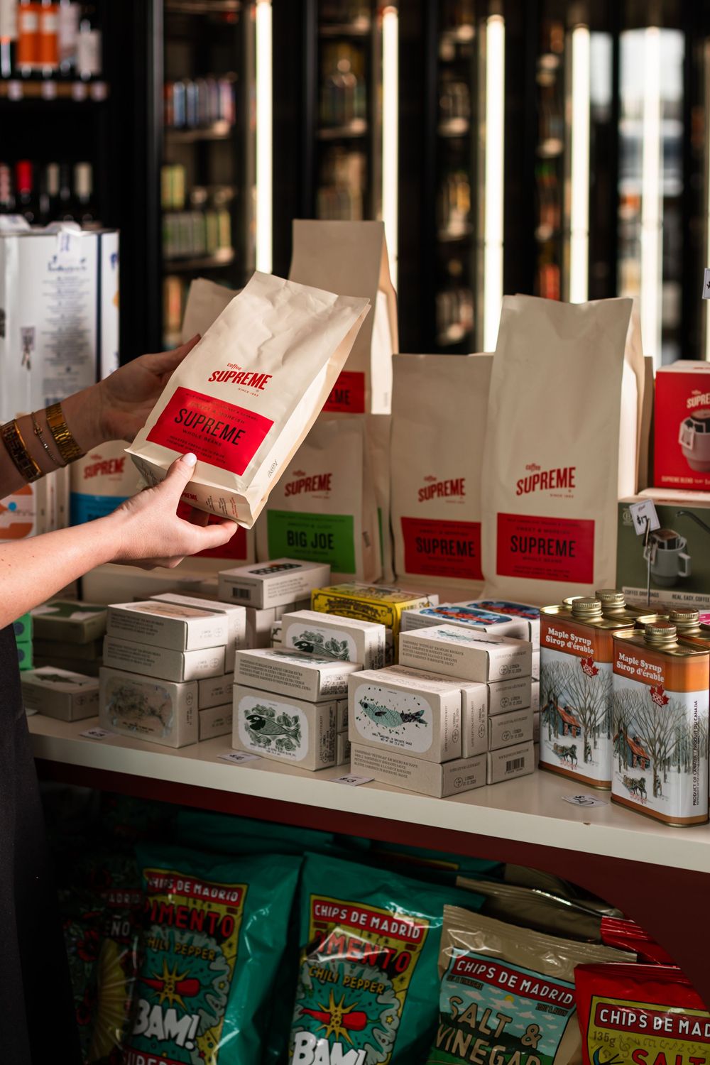 Hand holding a Supreme coffee bag with a red label above shelves of boxed goods, tinned products and Chips de Mais snack bags in a shop.