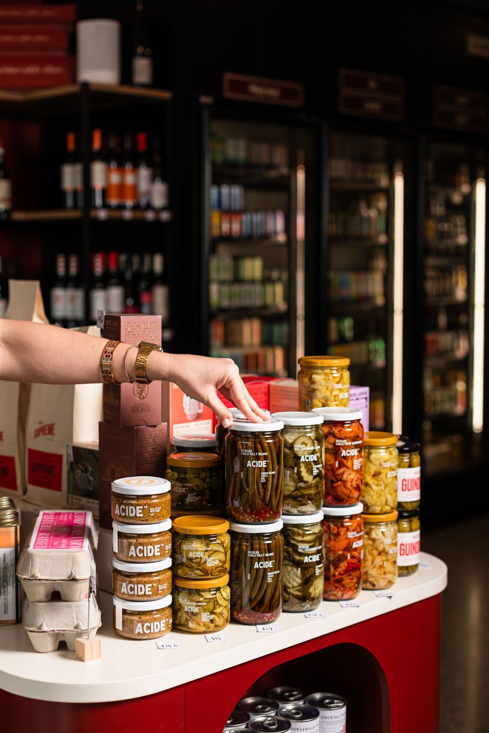 Hand reaching for jars of Acide pickled vegetables displayed on a red and white shop counter with wine bottles and shelves behind.