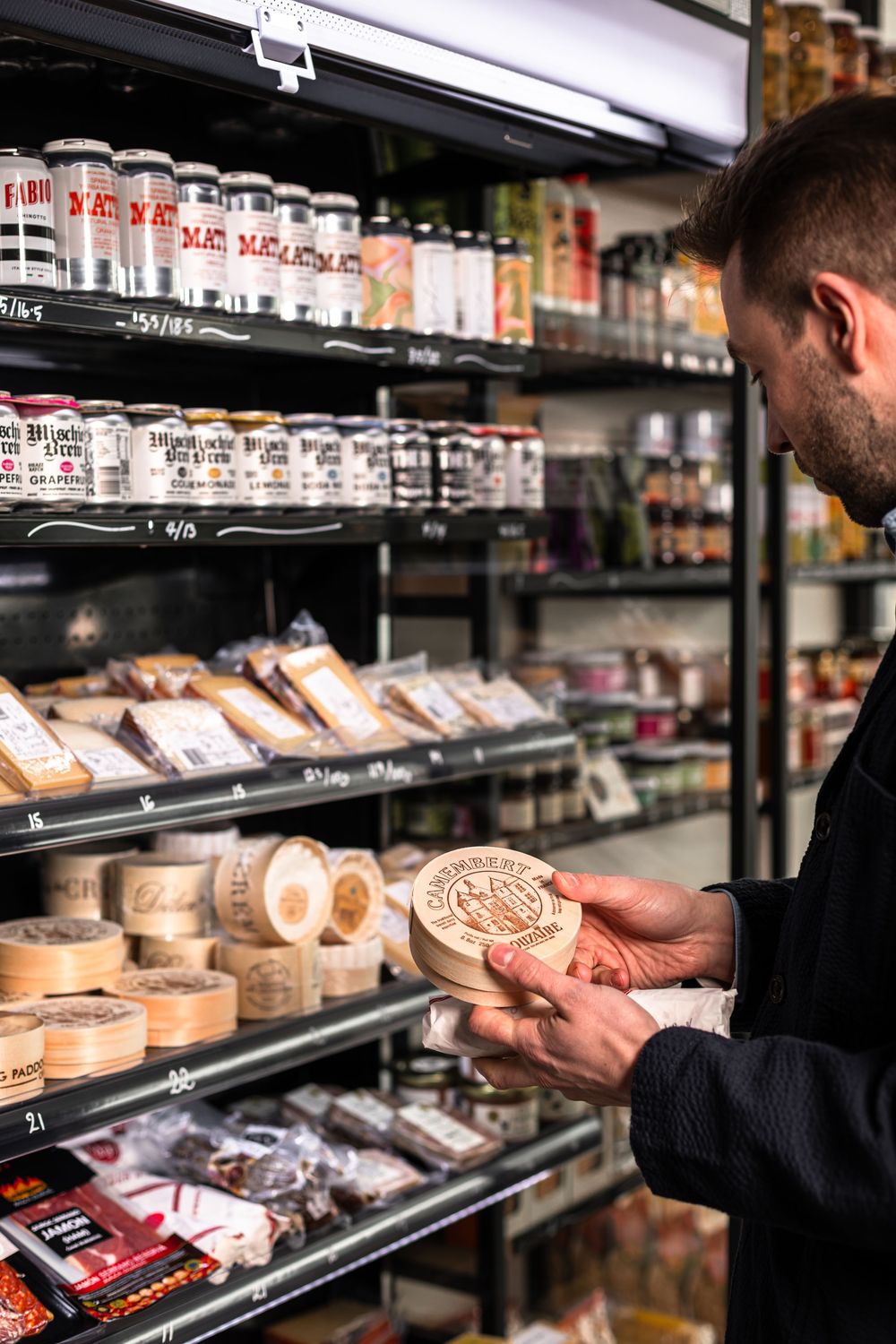 Shopper holding a round wooden box of cheese in a specialty food store aisle with shelves of canned goods, jars, and packaged deli items.