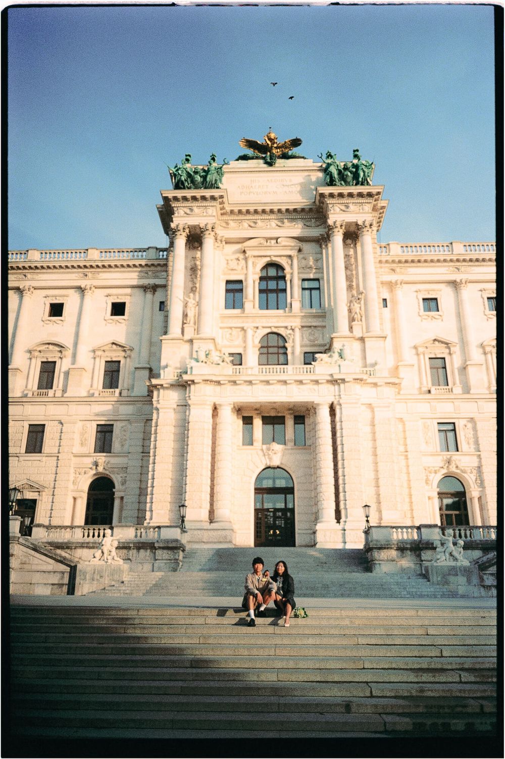 Two people sit on wide stone steps in front of a grand neoclassical palace with tall columns and rooftop statues under a blue sky.