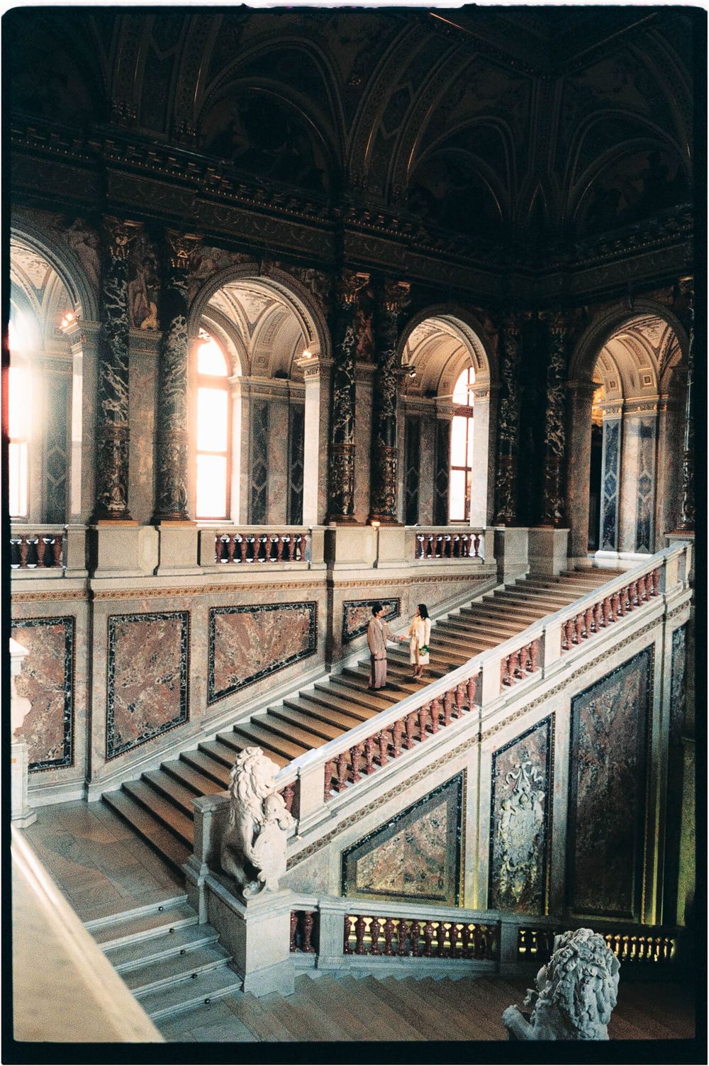 Two visitors climb the ornate marble grand staircase of a historic museum, framed by arched windows, carved balustrades and sculpted lions.
