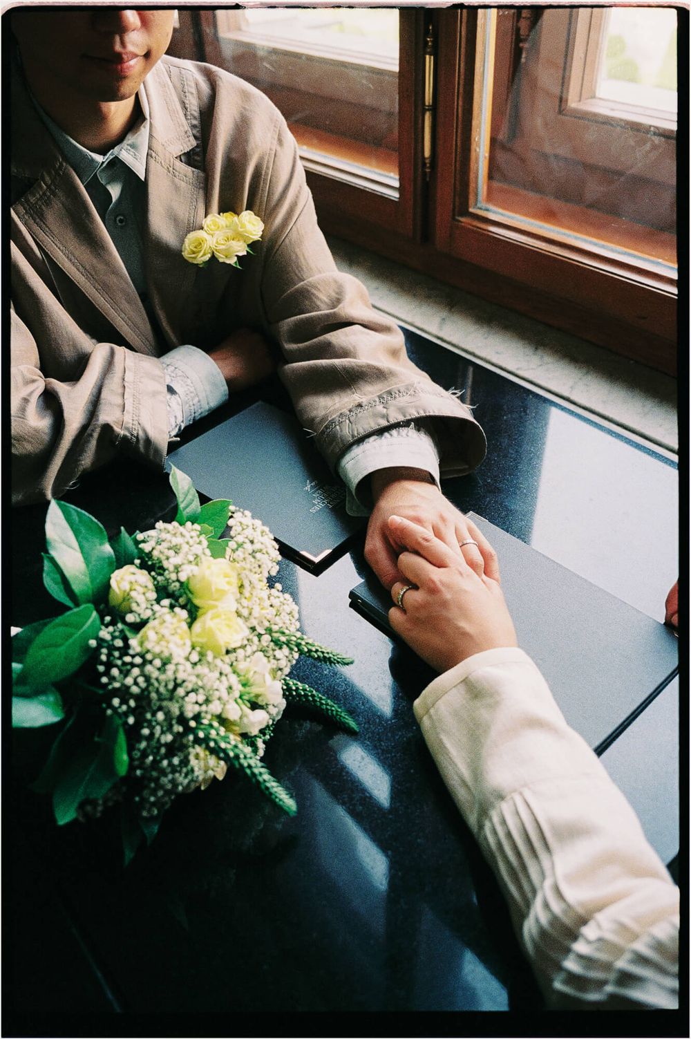 Groom with a yellow rose boutonniere holds the bride's hand beside a white and yellow wedding bouquet on a black table.