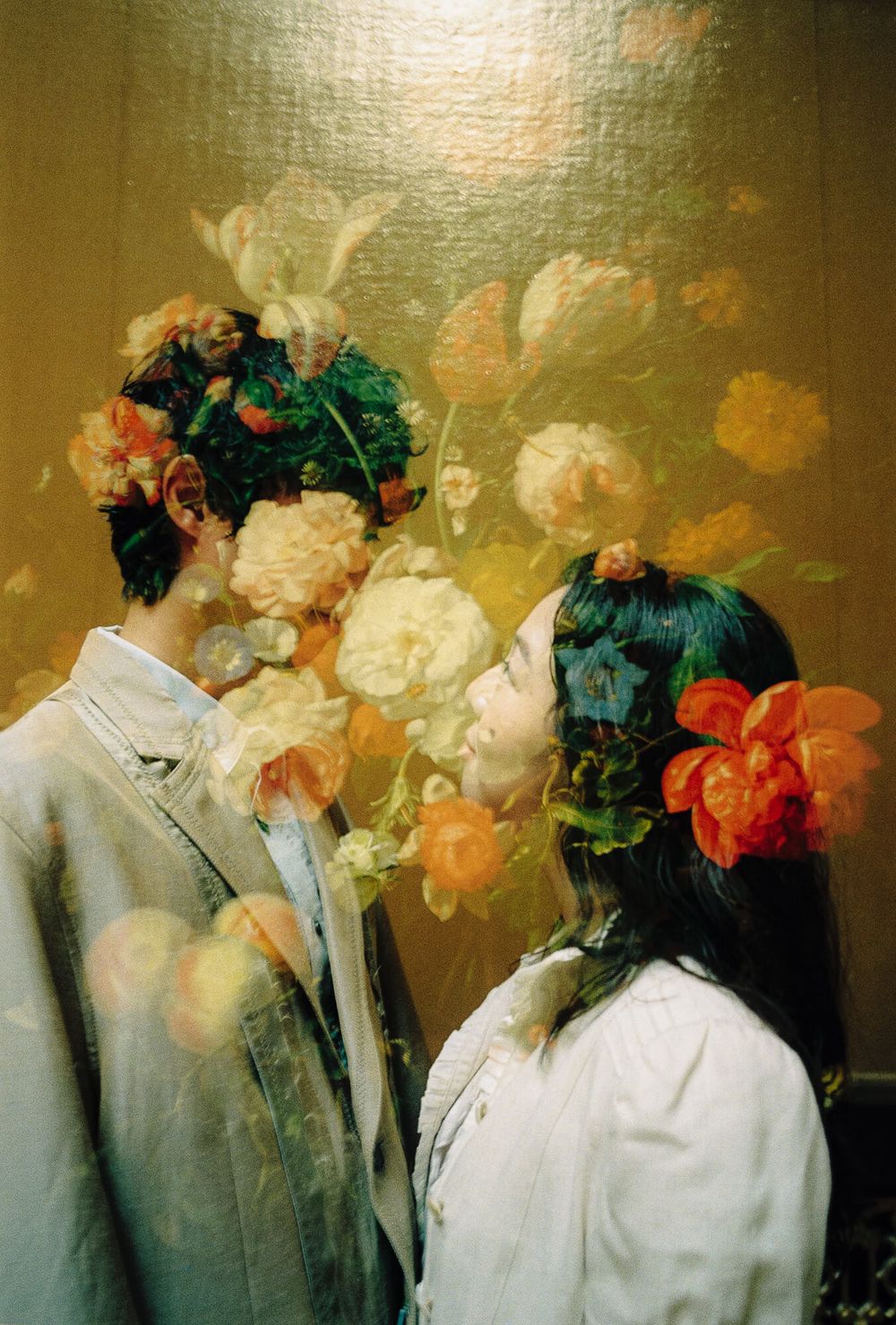 Two people face each other in profile against a golden floral backdrop, their hair and faces blended with orange and cream blooms.