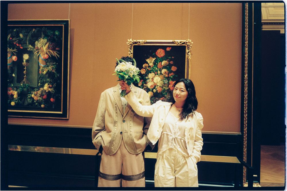 Two people in light trench coats stand in a museum gallery, one holding a bouquet over their face, before a floral still life painting.