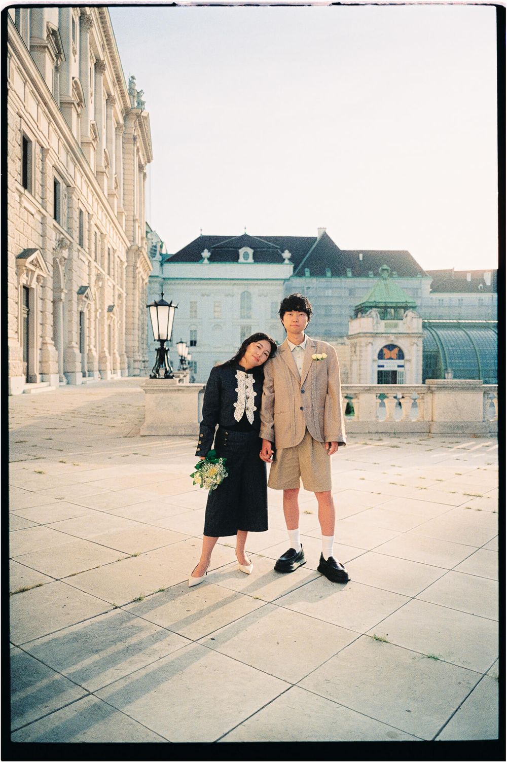 Couple in stylish outfits holding hands and posing on a sunlit stone plaza in front of historic European palace architecture.