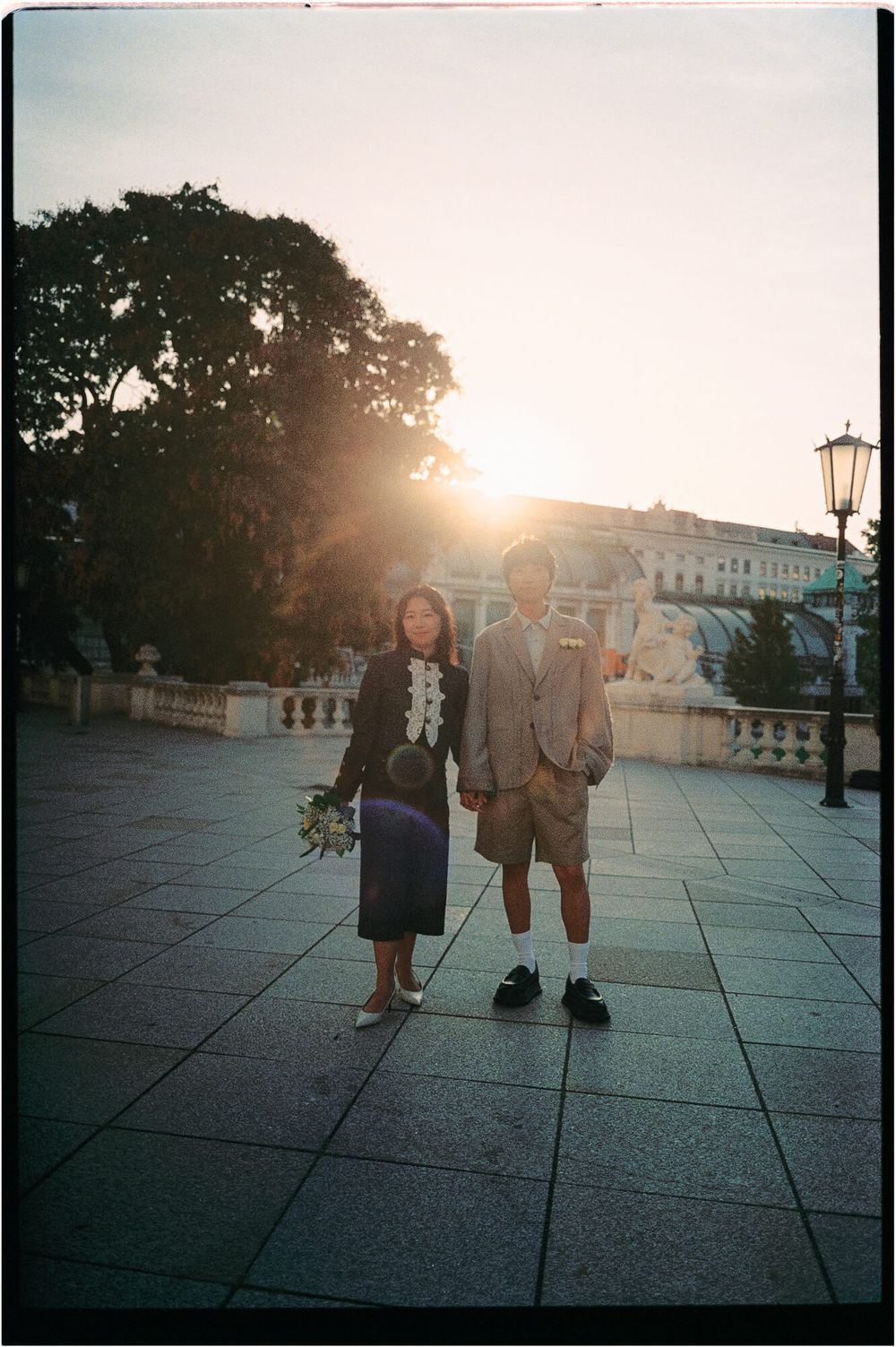 Couple holding hands and a bouquet on a stone plaza at sunset, with warm sunlight glowing behind trees and a historic building.
