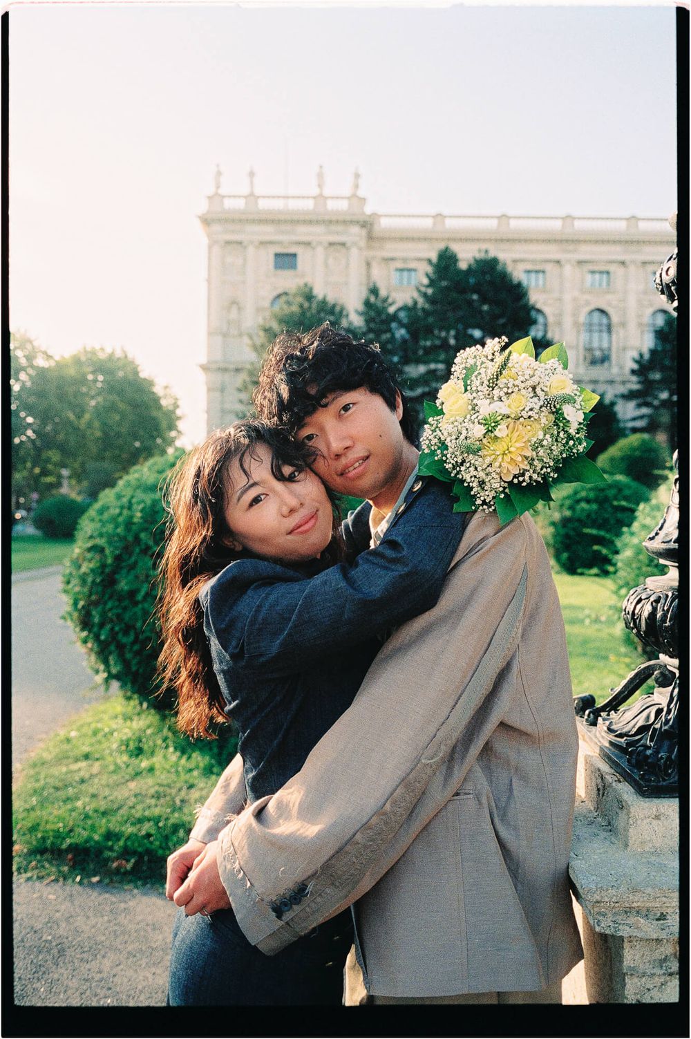 Couple embracing outdoors in a sunlit park, holding a bouquet of white and yellow flowers with a grand historic building behind them.