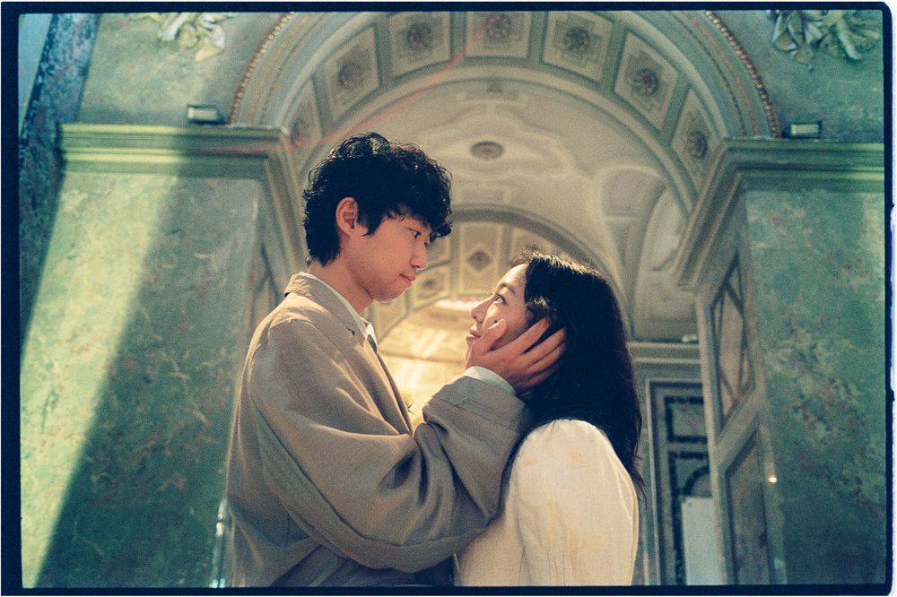 A man in a gray coat gently cups a woman's face as they stand close in an ornate arched hallway with green marble walls.