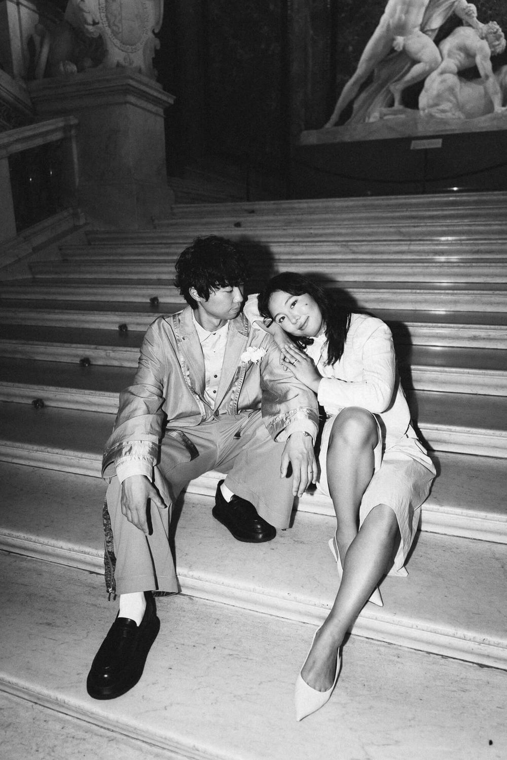 Black and white photo of a couple sitting close on marble museum steps, she rests her head on his shoulder near classical sculptures.