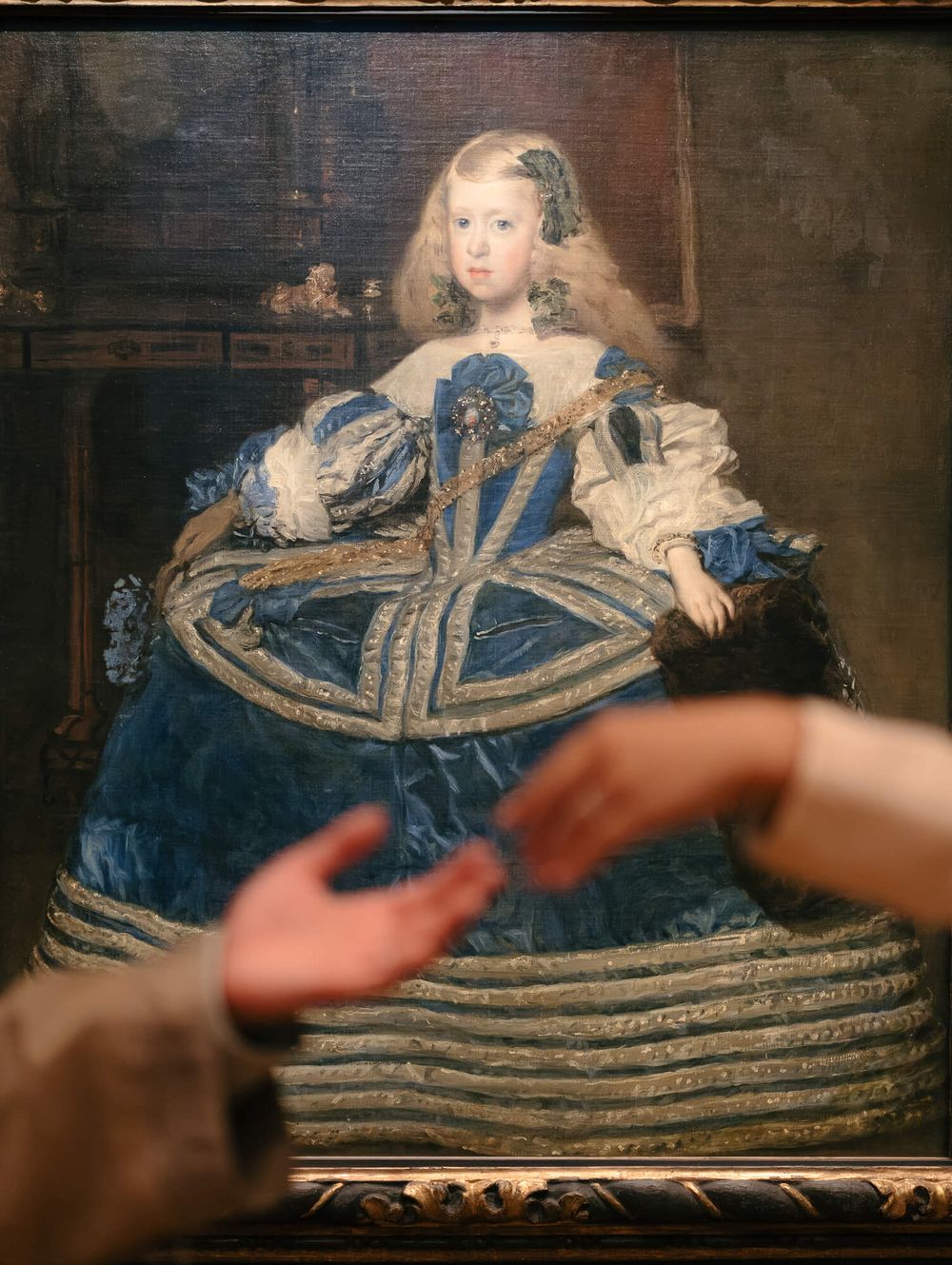 Baroque portrait of a young girl in a blue silk gown with silver trim and wide panniers, as two blurred hands gesture in front.
