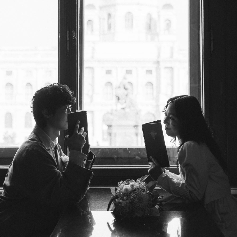 Black and white photo of a man and woman sitting across a cafe table reading books, with a flower bouquet and a grand building outside.