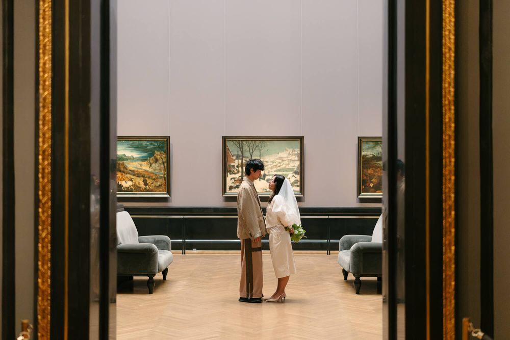 A bride and groom stand facing each other in a gallery hall with framed landscape paintings, gray armchairs and a warm wooden floor.