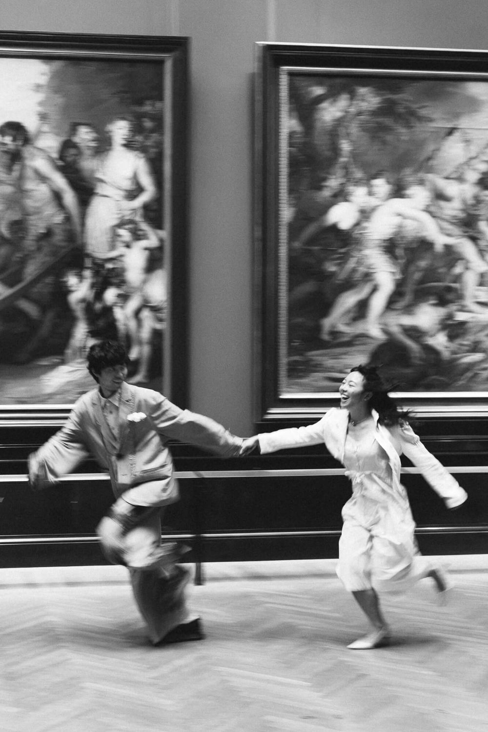 Two people in light clothing hold hands and run joyfully through an art museum gallery past large framed classical paintings.