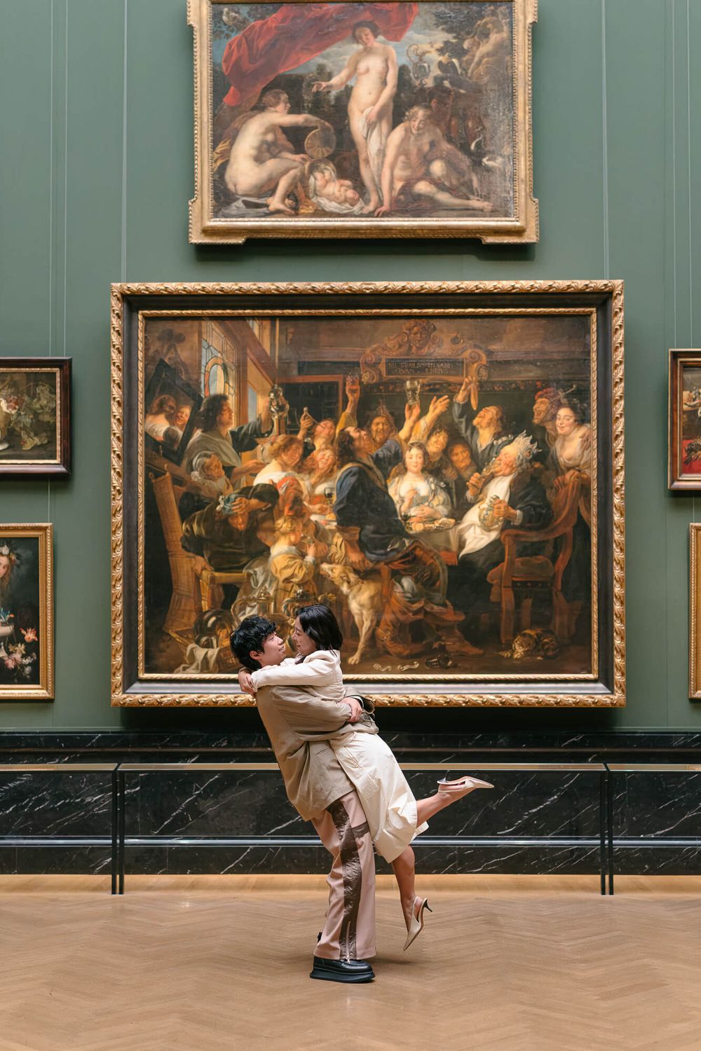 Couple embracing in a romantic lift in front of large Baroque paintings in a museum gallery with green walls and a herringbone wood floor.