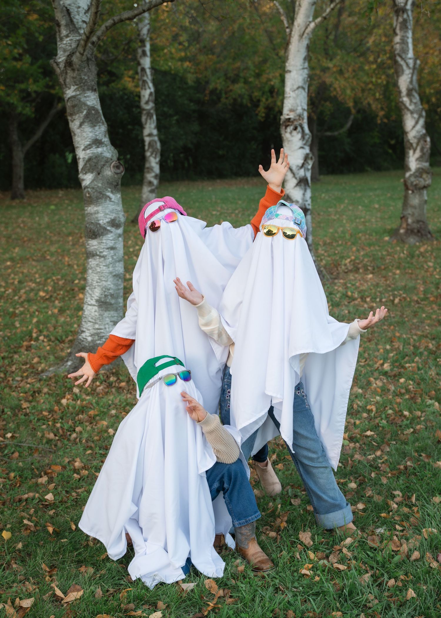 Three children in homemade white sheet ghost costumes with colorful hats and sunglasses pose with raised hands among birch trees in a park.