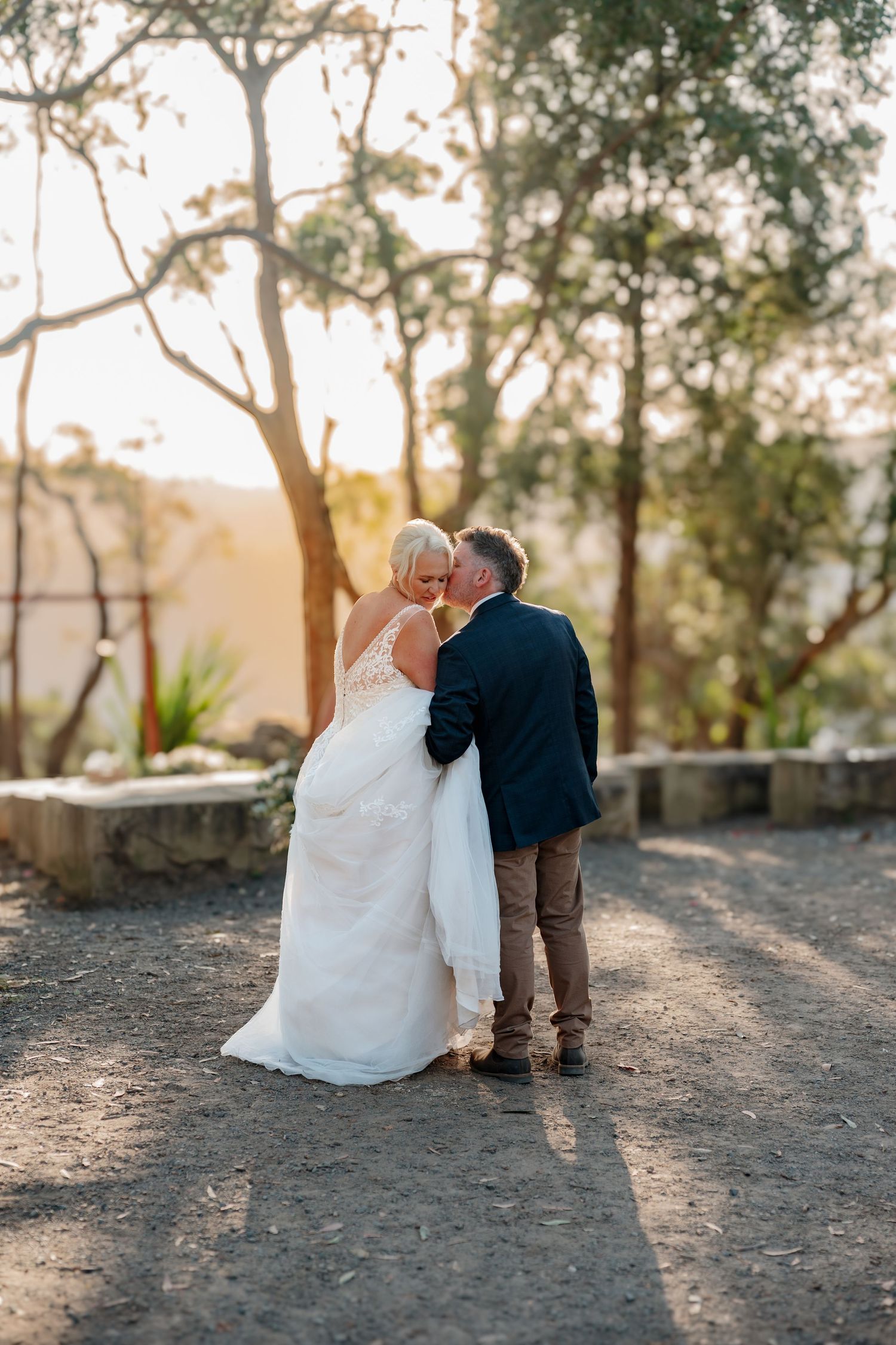 Bride and groom sharing a romantic kiss at golden hour outdoors, surrounded by trees in a scenic wedding setting.
