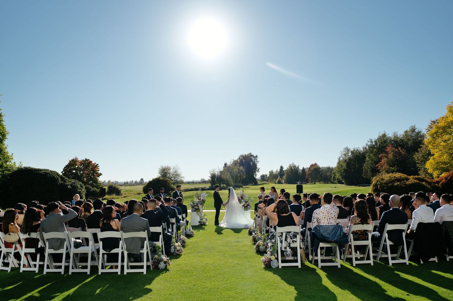 Wedding guests seated in white chairs on a sunny green lawn watch a bride in a white gown and groom exchange vows at an outdoor ceremony.