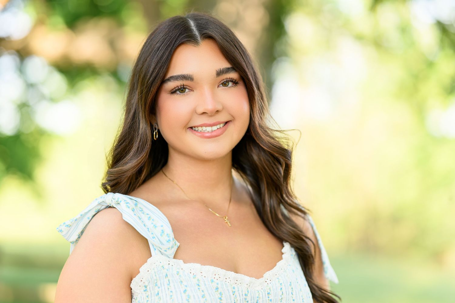 Smiling teenage girl with long brown hair wearing a light blue floral top, posing outdoors with green trees.