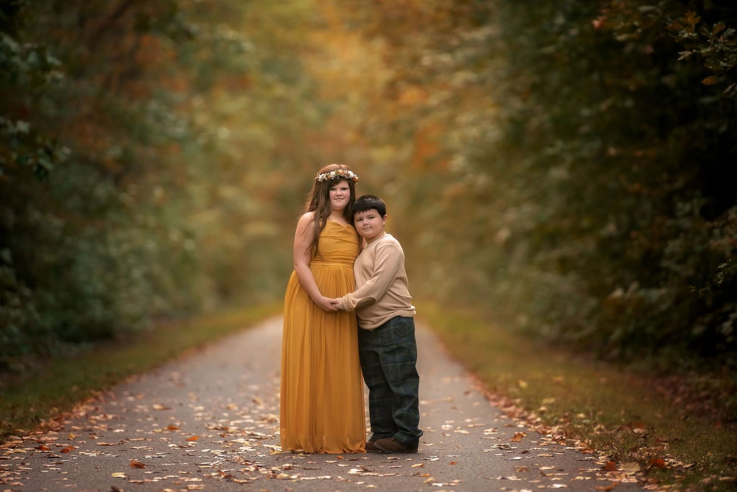 Woman in a flowing mustard maxi dress with a floral crown holds hands with a boy on a leaf-covered path in an autumn forest.