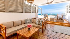 Coastal living room with beige outdoor sofa and wooden coffee table featuring ocean views from covered patio.
