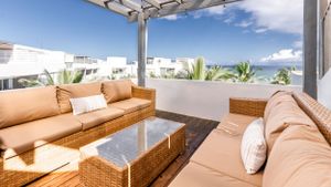 Modern rooftop lounge area featuring tan wicker furniture and glass table under a translucent pergola with ocean views.