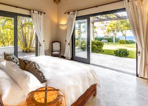 Luxurious bedroom with glass sliding doors opening to ocean view patio and white curtains framing tropical landscape.