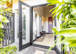Modern black-framed glass entrance doors surrounded by lush tropical plants and gravel pathway.