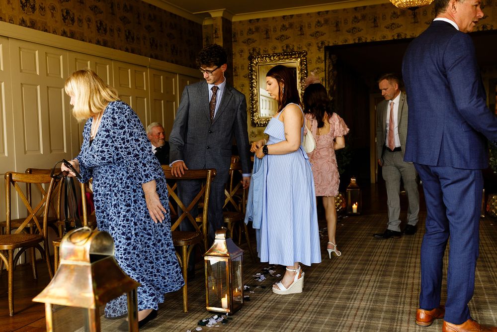 Elegantly dressed wedding guests mingle in a grand hallway with plaid carpet, wooden chairs, and decorative lanterns.