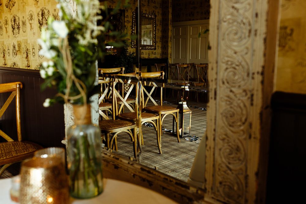 Elegant rustic venue with ornate mirror reflecting wooden cross-back chairs on vintage tile floors, perfect for weddings.