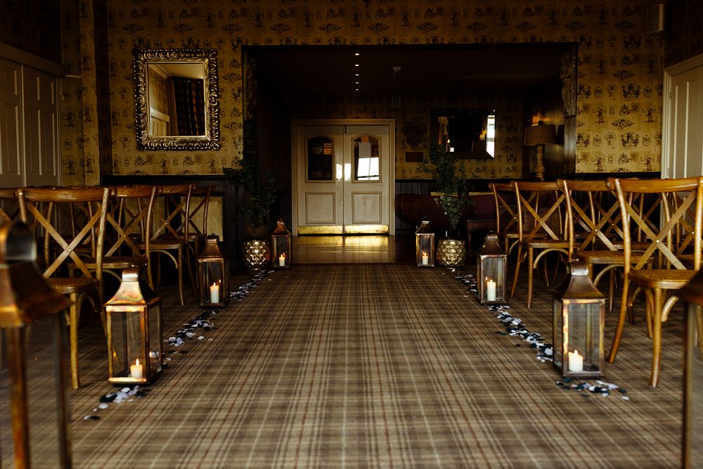 Elegant wedding ceremony aisle with wooden cross-back chairs, lanterns, and tartan carpet in a warmly lit vintage venue.