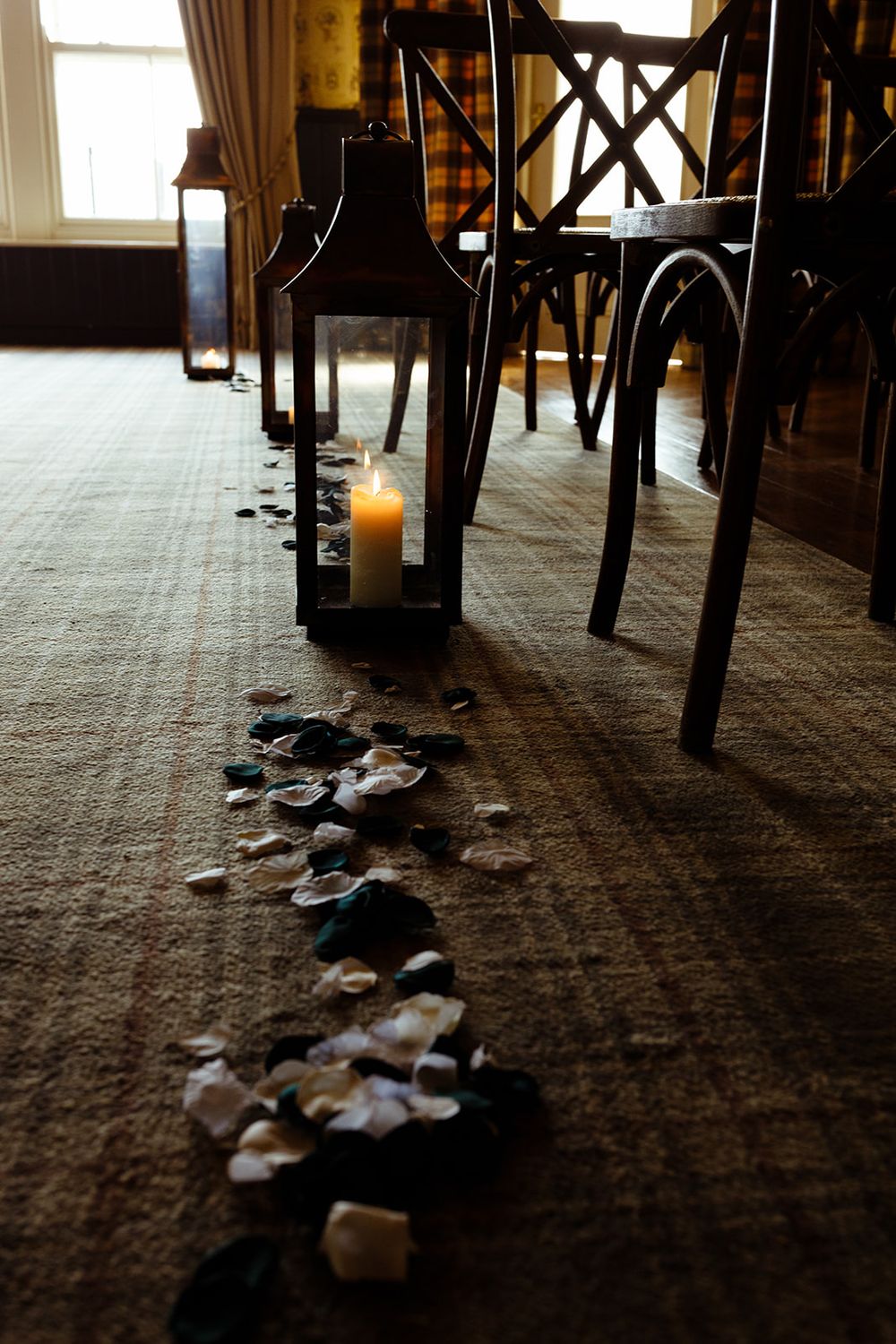 Elegant wedding aisle with rose petals, lit candle lantern, and dark wooden chairs on carpet.