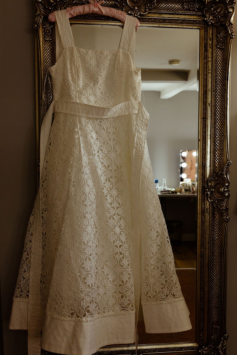 Elegant white lace wedding dress hanging on a gold ornate mirror in a bridal suite.