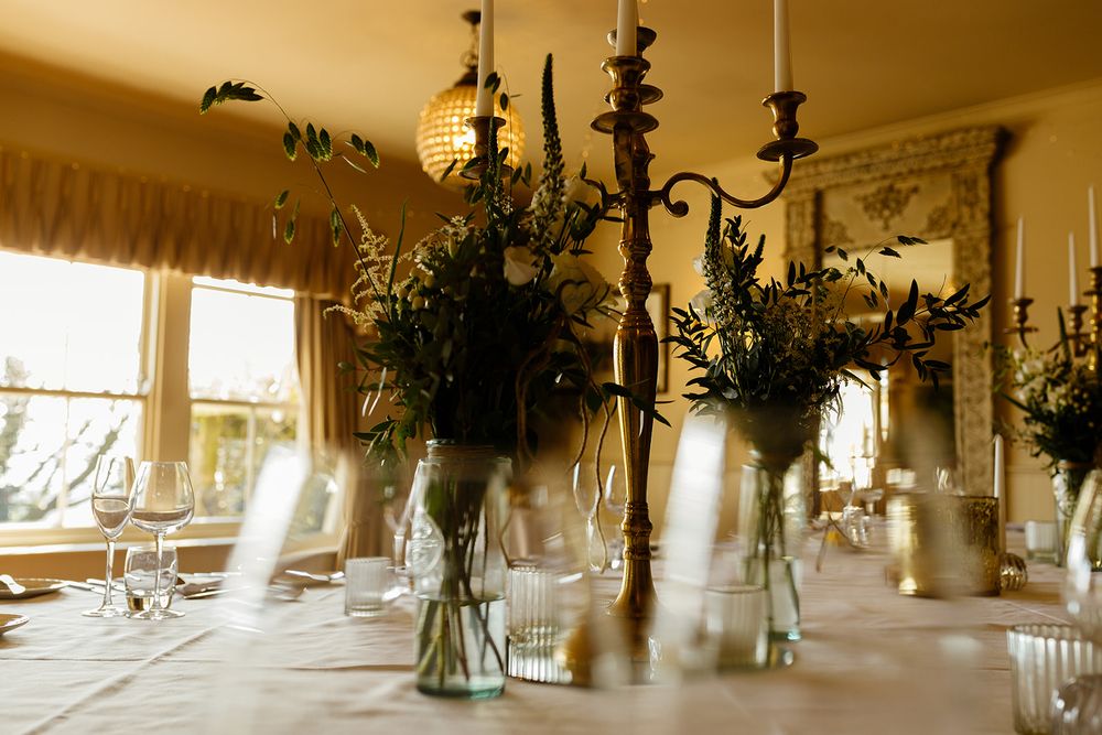 Elegant dining table with glass vases, greenery, and candelabra centerpieces in a warm, sunlit room.