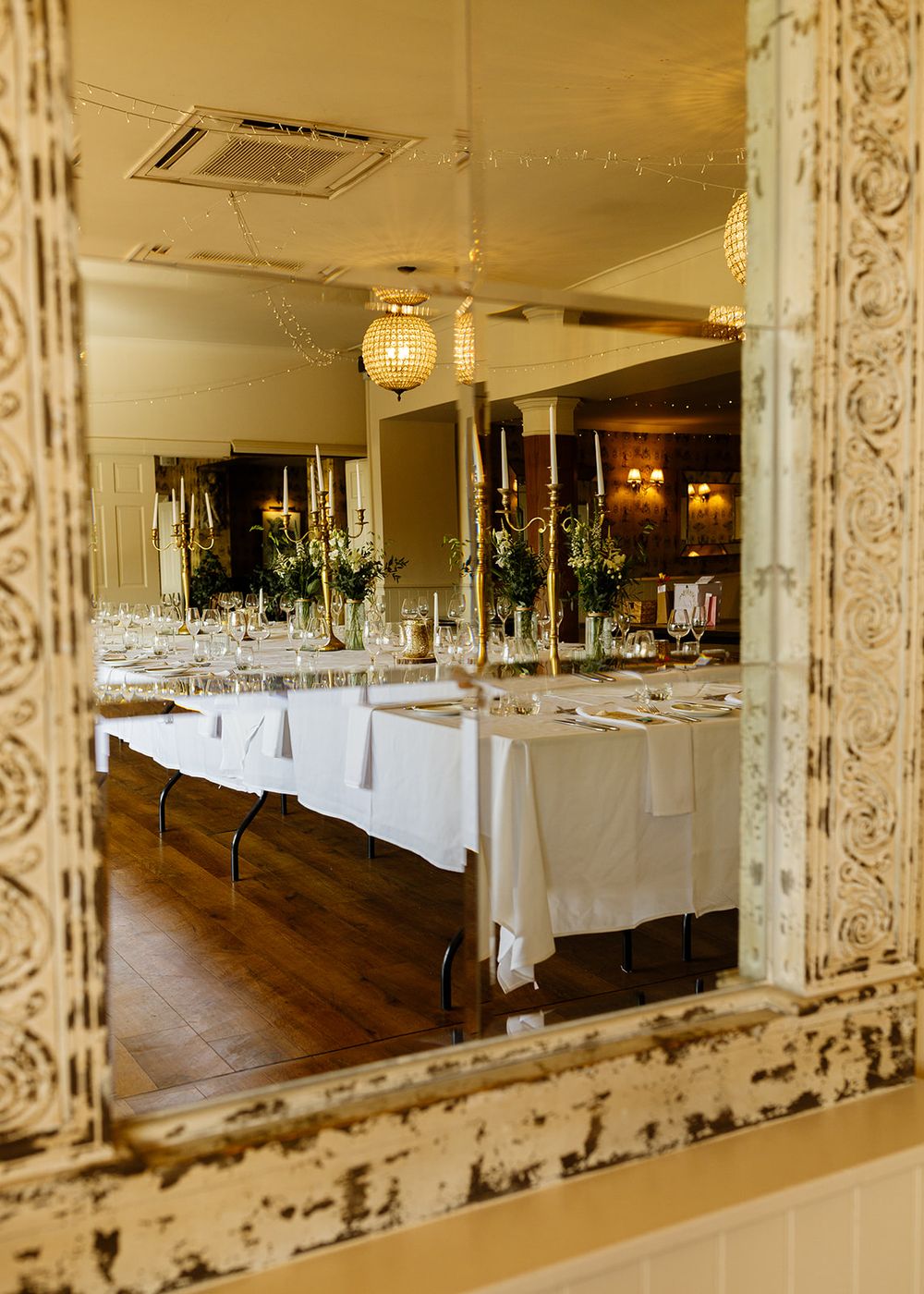 Elegant wedding reception dining room reflected in ornate distressed white mirror with long white tablecloths and gold candelabras.