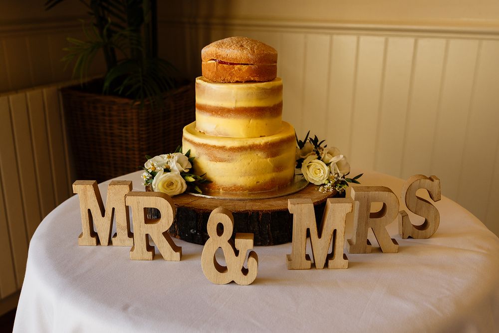 Elegant naked wedding cake on wooden slice with MR & MRS letters and white roses on a white tablecloth.