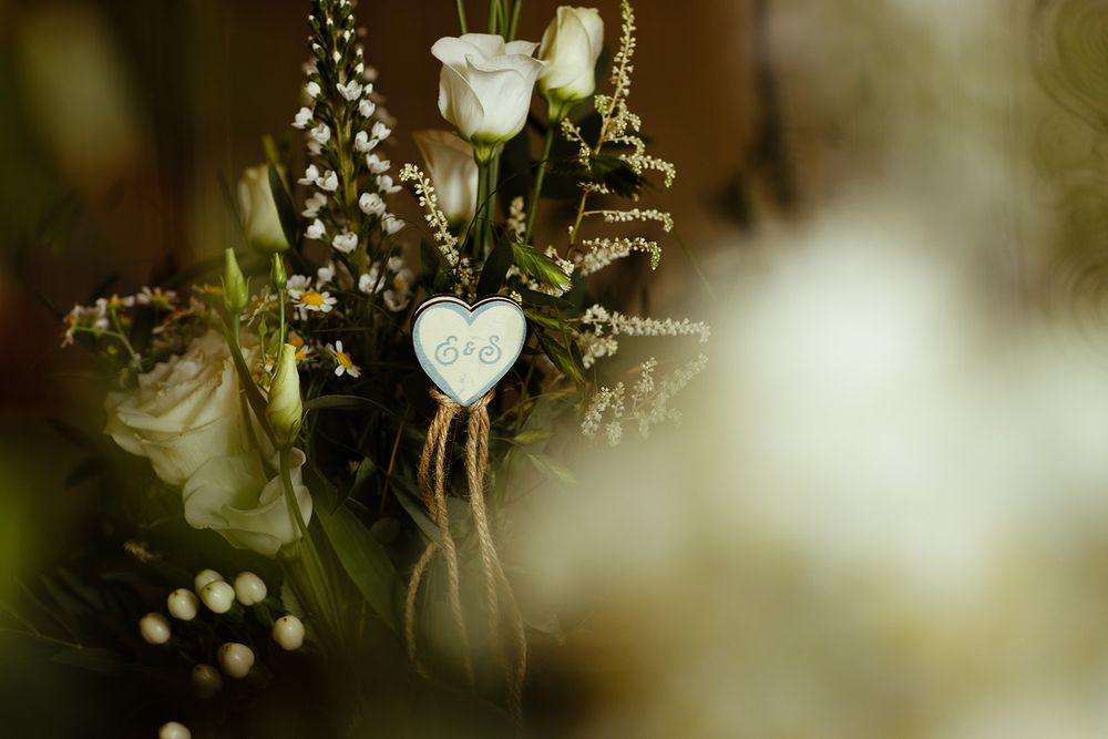 White roses and greenery bouquet with a rustic heart charm tied with twine, perfect for a romantic wedding arrangement.