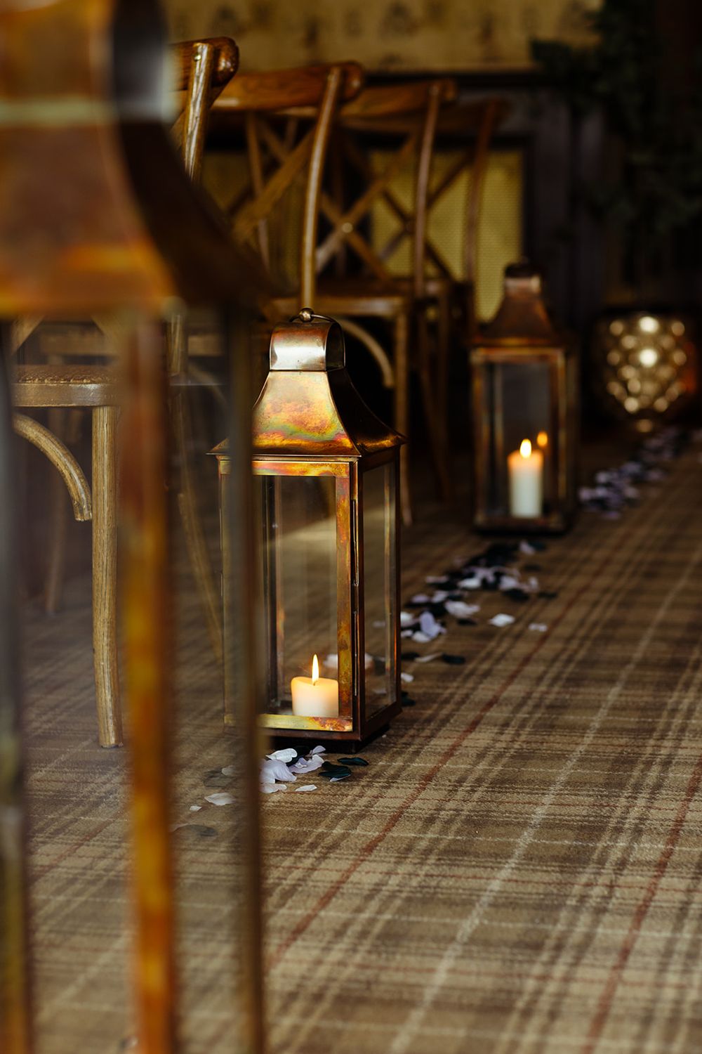 Rustic wedding aisle decorated with antique brass lanterns holding lit candles on a plaid carpet beside wooden chairs.